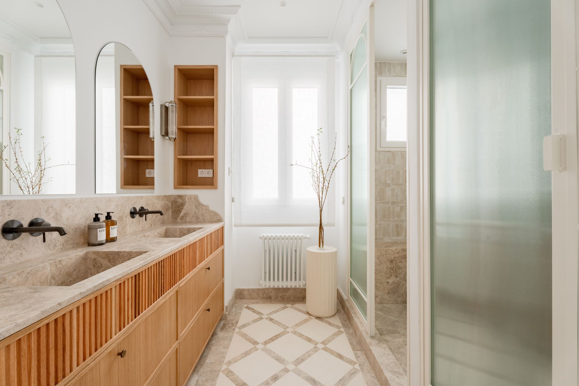 Baño luminoso con mueble de madera acanalada, lavabo doble de piedra, espejos en arco y mampara de vidrio acanalado