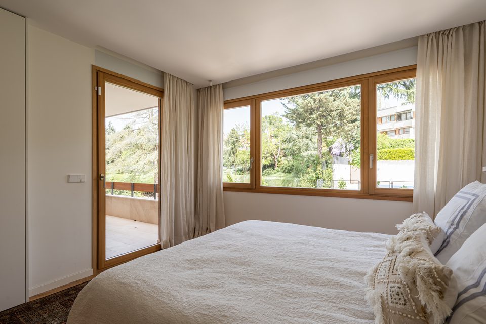 Dormitorio con ventana y balconera de madera, cortinas de lino y vistas a un jardín