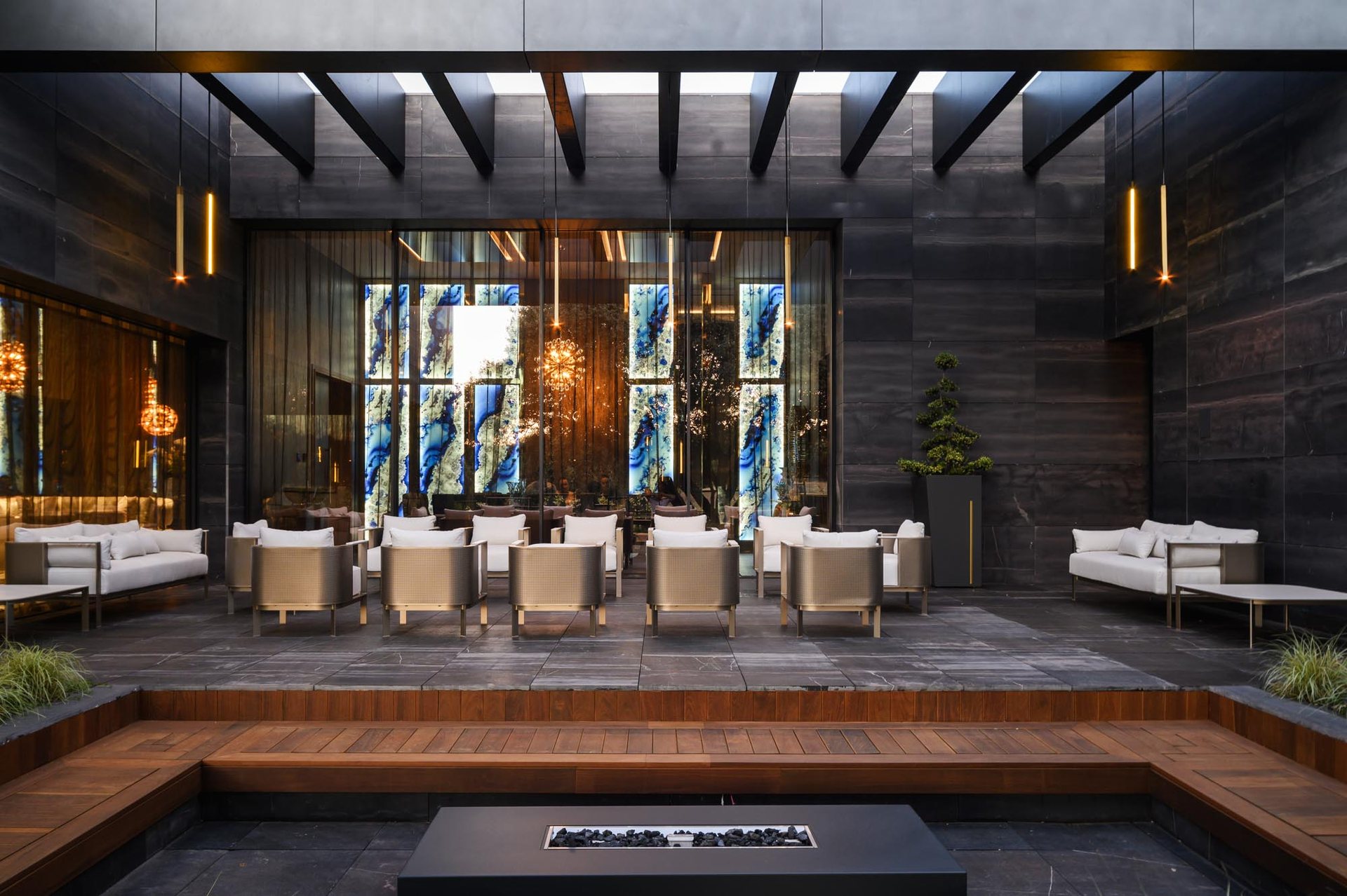 Patio-lounge de un hotel de lujo con sillones de latón y cojines blancos, muros de piedra oscura, paneles de ónix retroiluminados y banco de madera junto a una mesa de fuego